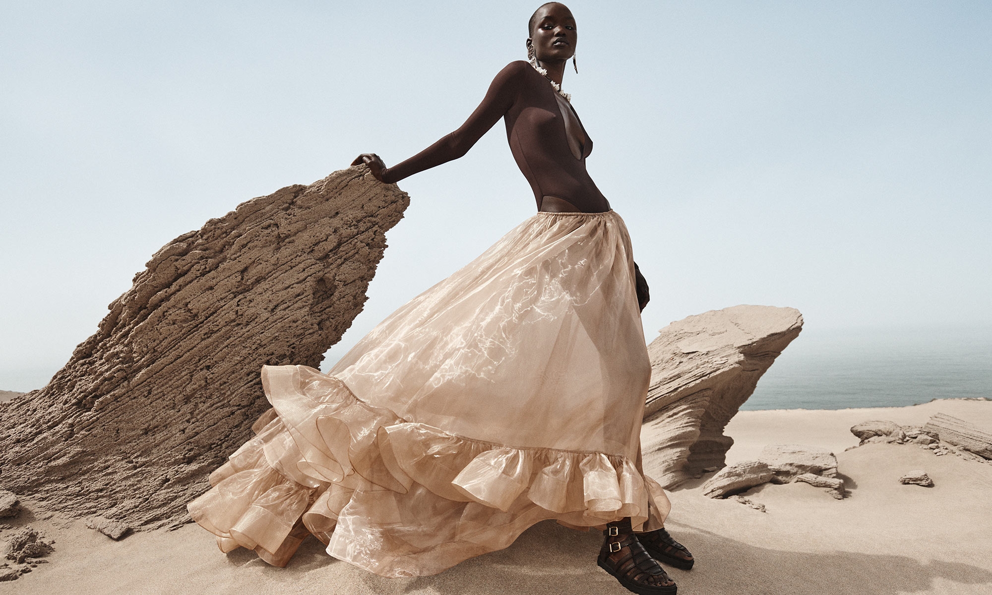 Model is wearing the Dawning Scoop Bodysuit in Chocolate, Dawning Flounce Maxi Skirt in Gold, Goldie Gladiator in Espresso, Ore Chandeliers in Gold/Copper and Mellow Pearl Beaded Necklace in Gold/Mult 